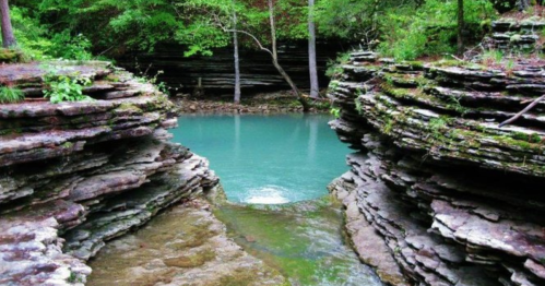 A serene blue pool surrounded by layered rock formations and lush greenery in a tranquil natural setting.