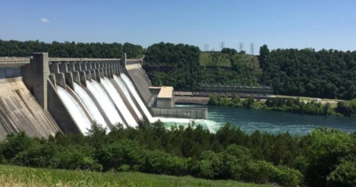 A large dam with water flowing from its gates, surrounded by green trees and a clear blue sky.