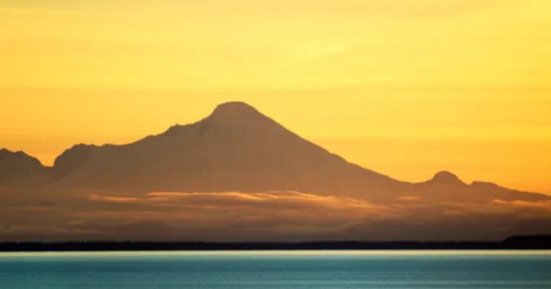 A silhouette of a mountain against a vibrant sunset sky, reflecting on calm waters below.
