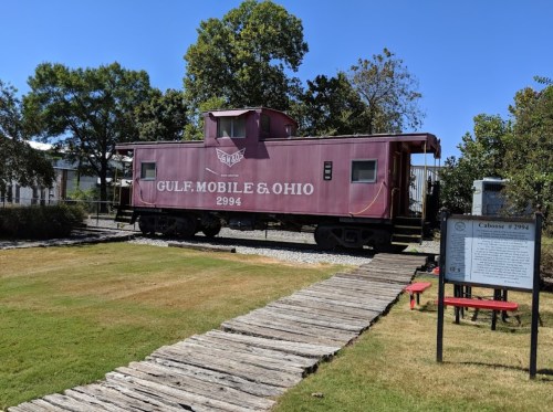 A vintage Gulf, Mobile & Ohio train caboose displayed on a grassy area with a wooden walkway and informational sign nearby.