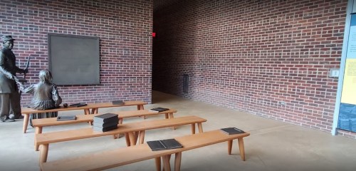 A classroom scene with wooden benches, a chalkboard, and a statue of a teacher and student against a brick wall.