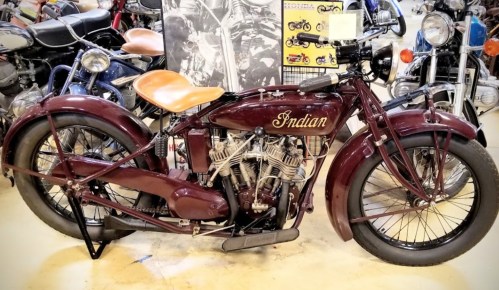 A vintage Indian motorcycle with a maroon body and brown leather seat, displayed among other classic bikes.