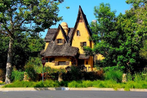 A whimsical yellow house with a steep, pointed roof, surrounded by lush greenery and trees on a sunny day.