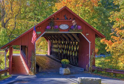 A red covered bridge adorned with flowers, surrounded by vibrant autumn foliage and an American flag.