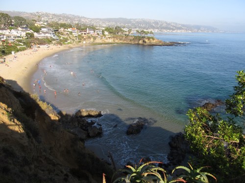 A scenic beach view with people enjoying the water, surrounded by cliffs and houses under a clear blue sky.