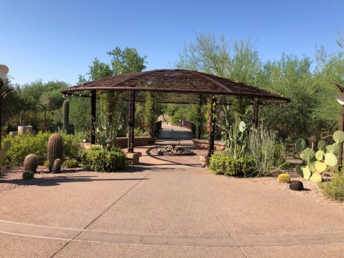 A pathway leads through a desert garden with cacti and a shaded gazebo under a clear blue sky.