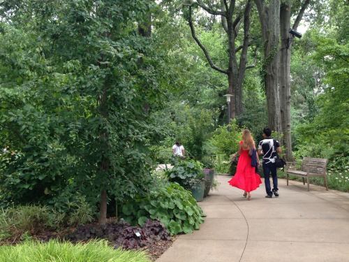 A woman in a red dress walks along a path in a lush garden, accompanied by a person in a black and white outfit.