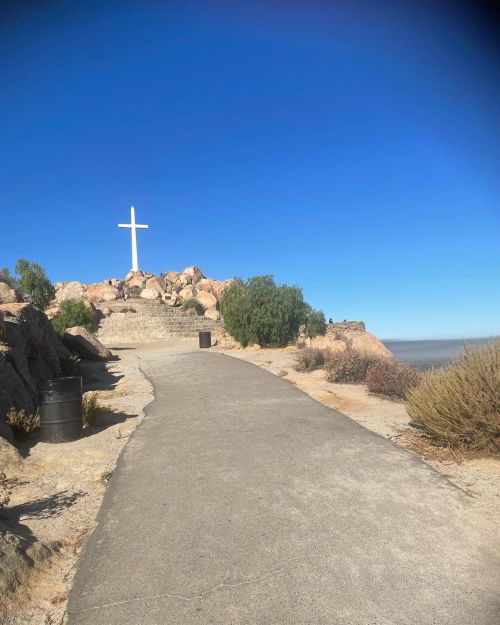 A pathway leads to a large white cross atop a rocky hill, surrounded by shrubs and a clear blue sky.