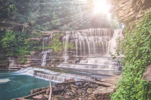 A serene waterfall cascades over rocky ledges, surrounded by lush greenery and illuminated by soft sunlight.