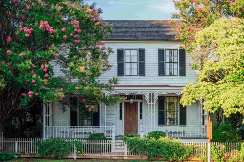 A charming two-story house with a porch, surrounded by blooming trees and a white picket fence.