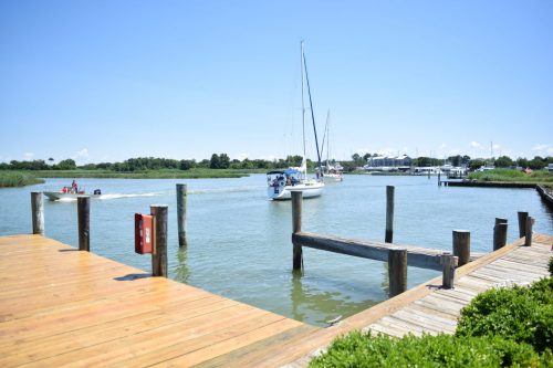 A calm waterway with a sailboat and dock, surrounded by greenery under a clear blue sky.