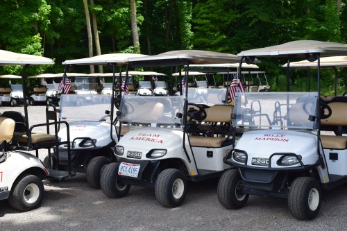 A row of golf carts parked on gravel, each with flags and personalized names on the front. Green trees in the background.