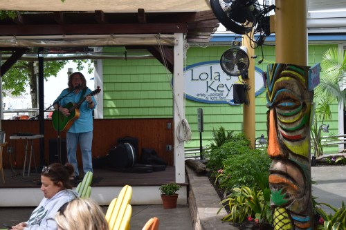 A musician plays guitar on stage at Lola's Key, with a colorful tiki statue and seated audience in the foreground.