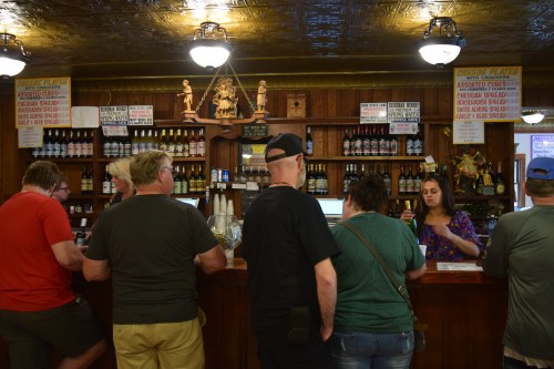 A group of people at a rustic bar, chatting with a bartender surrounded by shelves of bottles and menu signs.