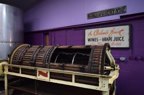 A vintage wine press in a purple room, with a sign reading "Bonded Winery No. 112" and "Wines & Grape Juice."