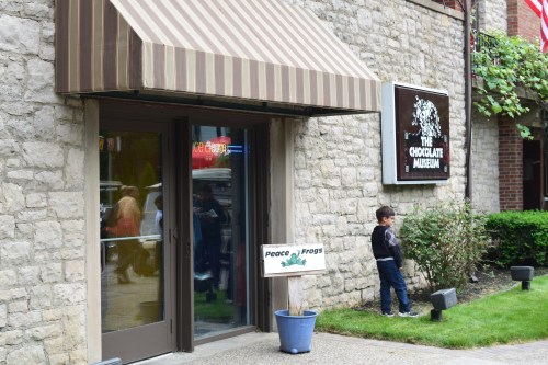 A child stands outside The Chocolate Museum, next to a sign for "Peace & Frogs," with a stone building in the background.