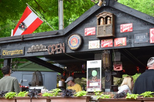 A rustic outdoor beer garden with an "Open" sign, flags, and patrons enjoying the atmosphere.