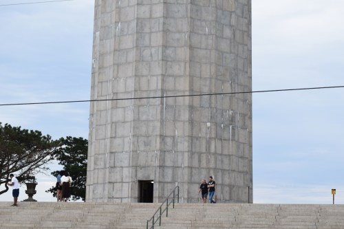 A tall stone tower with people walking nearby, set against a cloudy sky and trees in the background.
