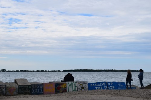 Two people stand by a lake, while a couple sits on a graffiti-covered barrier under a cloudy sky.