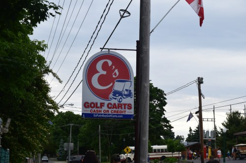 Sign for E's Golf Carts, offering cash or credit, with a cartoon golf cart graphic. Trees and power lines in the background.