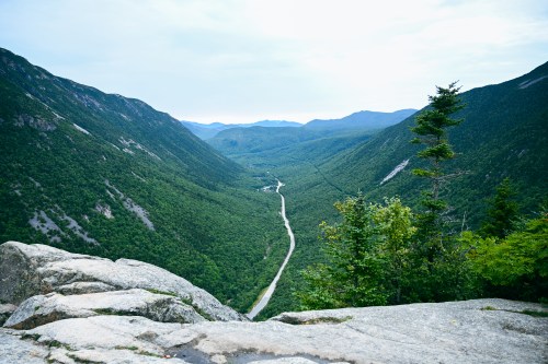 A panoramic view of a lush green valley with a winding road, framed by mountains under a cloudy sky.
