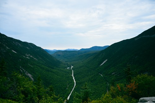 A scenic view of a lush green valley surrounded by mountains under a cloudy sky. A winding road cuts through the landscape.
