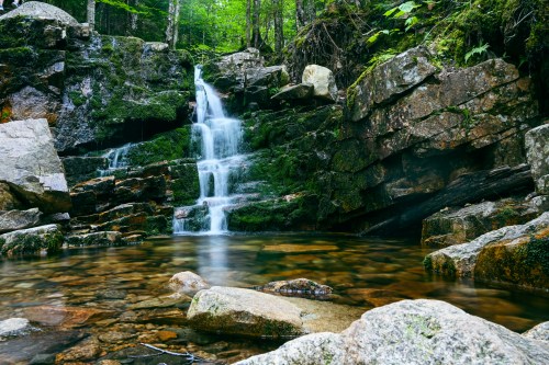 A serene waterfall cascades over rocks into a clear pool, surrounded by lush greenery and moss-covered stones.