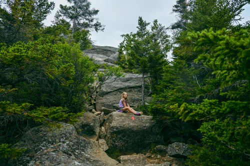 A girl sits on a large rock surrounded by lush green trees and rocky terrain in a natural setting.