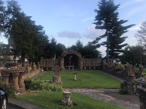 A stone garden with a central archway, surrounded by trees and decorative stone structures, under a blue sky.