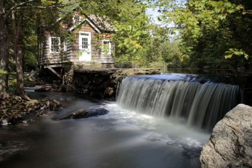 A serene scene featuring a small wooden house beside a flowing waterfall, surrounded by lush greenery.