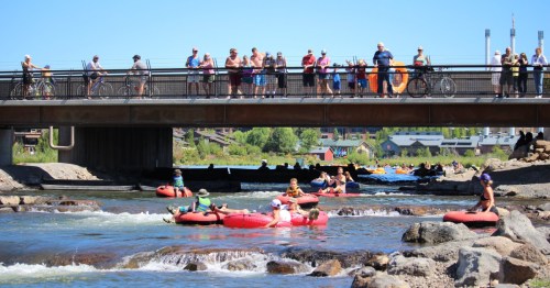 People enjoy tubing on a river while spectators watch from a bridge above on a sunny day.