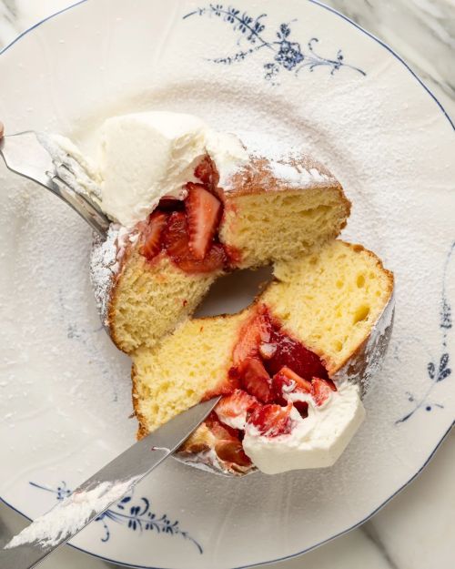 A sliced doughnut filled with strawberries and cream, dusted with powdered sugar, on a decorative plate.