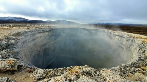 A large geothermal crater with steam rising, surrounded by rocky terrain and a cloudy sky in the background.