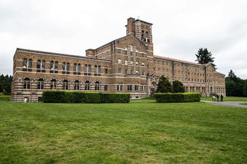 Historic brick building with a tower, surrounded by green grass and trees under a cloudy sky.
