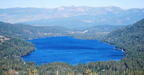 A serene blue lake surrounded by lush green forests and distant mountains under a clear blue sky.