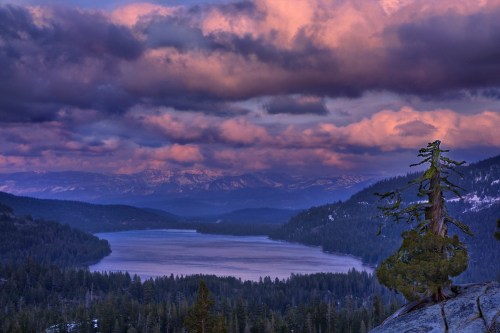 A serene lake surrounded by mountains under a dramatic sky with colorful clouds at sunset.