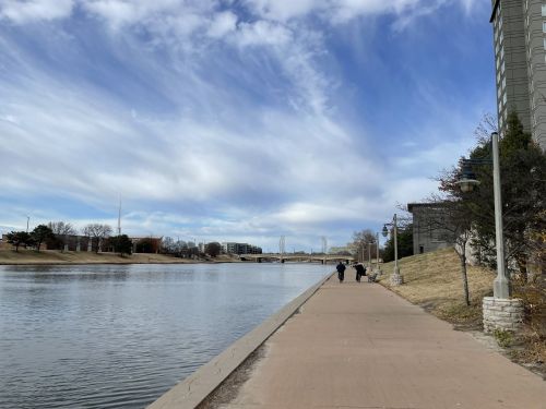 A scenic riverside path with two people walking, surrounded by trees and buildings under a cloudy sky.