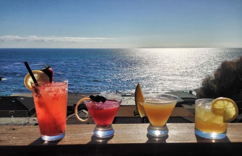Four colorful cocktails sit on a railing, overlooking a sparkling ocean under a clear blue sky.