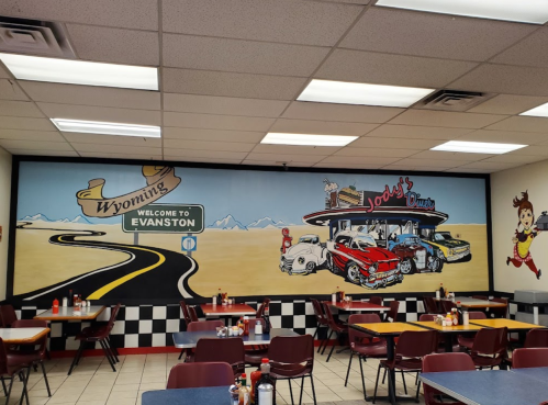 A diner interior featuring a mural of a road sign for Wyoming and classic cars, with empty tables and chairs.