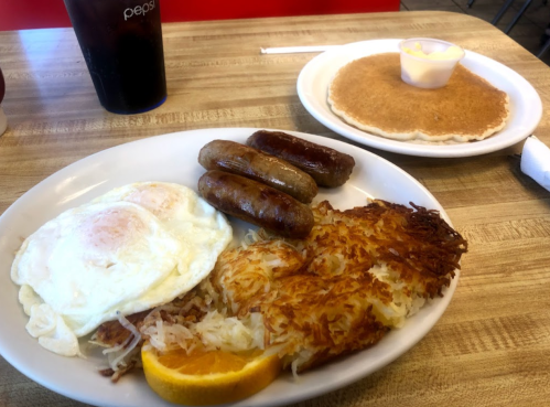 A plate with two fried eggs, sausage links, hash browns, and an orange slice, alongside a pancake with butter.