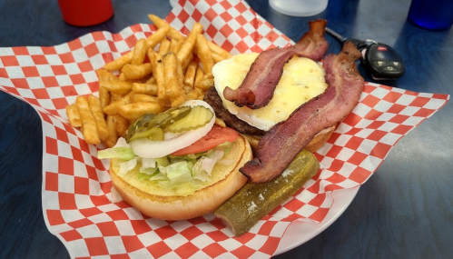 A plate with a burger topped with bacon and an egg, served with fries, pickles, and a side of lettuce and tomato.