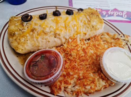 A plate featuring a large burrito topped with cheese and olives, accompanied by hash browns and two dipping sauces.