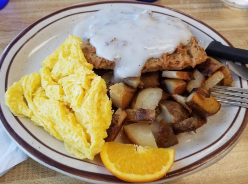 A plate of breakfast featuring scrambled eggs, fried potatoes, a biscuit with gravy, and a slice of orange.