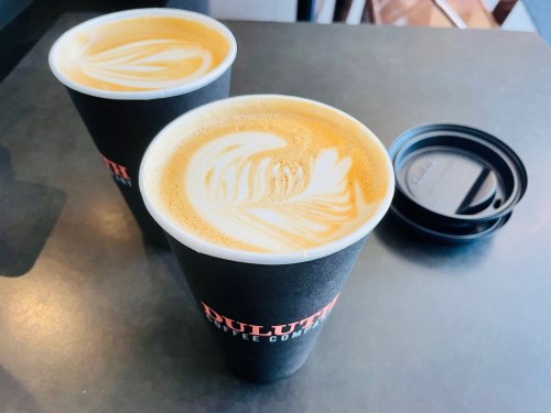 Two coffee cups with latte art on a dark table, one cup has a lid beside it.