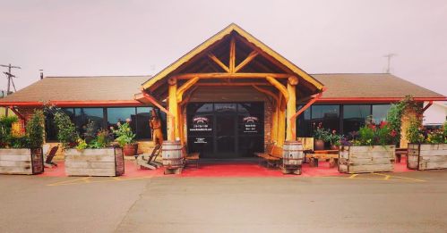 A rustic building with a wooden entrance, surrounded by flower planters and barrels, set against a cloudy sky.