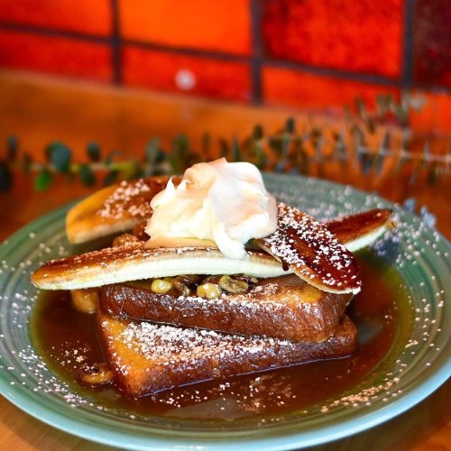 A plate of French toast topped with banana slices, whipped cream, and syrup, garnished with powdered sugar.