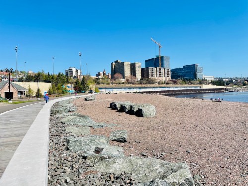 A sunny beach with large rocks, a boardwalk, and a city skyline in the background under a clear blue sky.