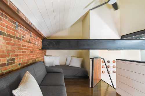 Cozy loft space with a gray sofa, white pillows, exposed brick wall, and wooden beams. Bright and inviting atmosphere.