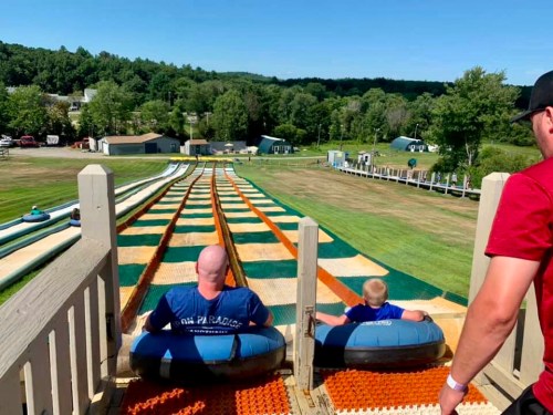 Two people sit on inflatable tubes at the top of a large water slide, ready to descend into a grassy area below.