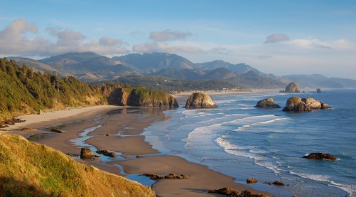 A scenic beach view with gentle waves, rocky formations, and mountains in the background under a clear blue sky.